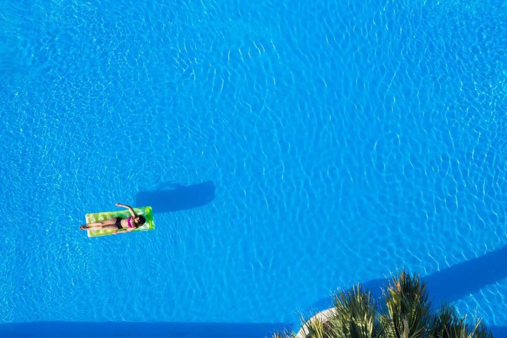 Sieviete guļ uz zaļas pludiņa, baudot sauli skaidrā zilā baseinā. Ūdens virsma spīd un veido viļņus, radot relaksējošu atmosfēru.