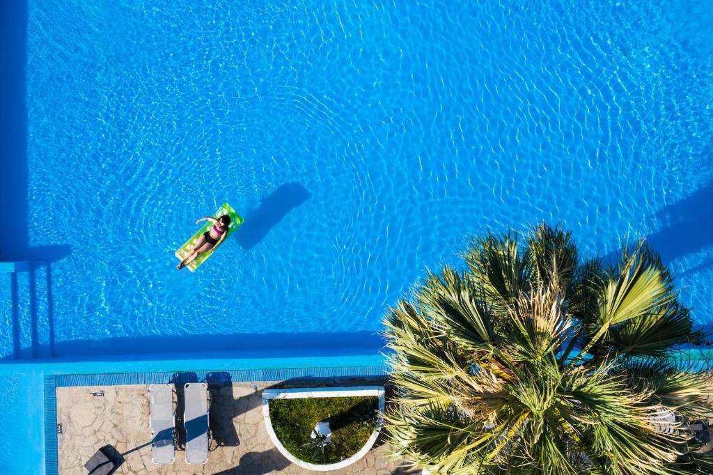 Peldbaseins ar spilgtu zilu ūdeni, kurā atpūšas cilvēks uz peldēšanas dēļa. Blakus ir palmas un sauļošanās krēsli, radot relaksējošu atmosfēru.