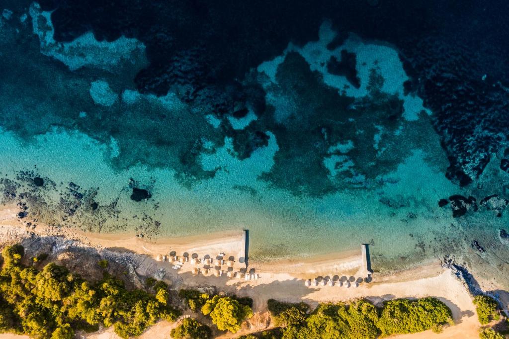 Skaists gaisa skats uz pludmali ar smilšu krastu un skaidru zilo ūdeni, kur redzami sauļošanās krēsli un koka piestātne. Ideāla vieta atpūtai un relaksācijai.