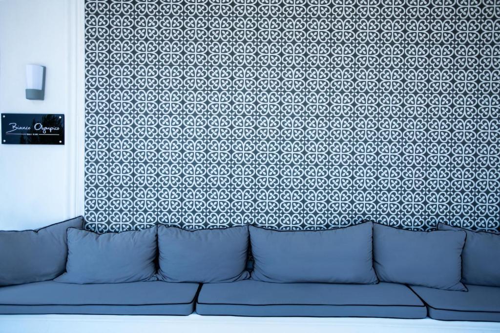 Mūsdienīgs dīvāns ar pelēkiem spilveniem, kas izvietots pretī dekoratīvai sienai ar rakstainu dizainu. Sienā ir arī nosaukuma plāksne "Bianco Olimpico".