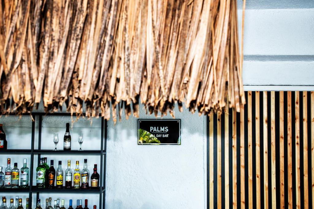 Trokšņainā bāra interjers ar palmu lapām un alkoholisko dzērienu plauktu. Redzams "PALMS DAY BAR" zīme.