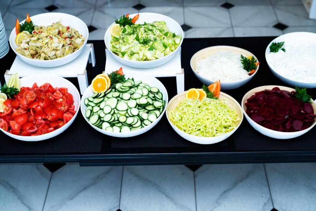 Dažādas salātu bļodas ar svaigiem dārzeņiem, tostarp tomātiem, gurķiem, kāpostiem un biešu salātiem, dekorētas ar citrusaugļiem un zaļumiem.