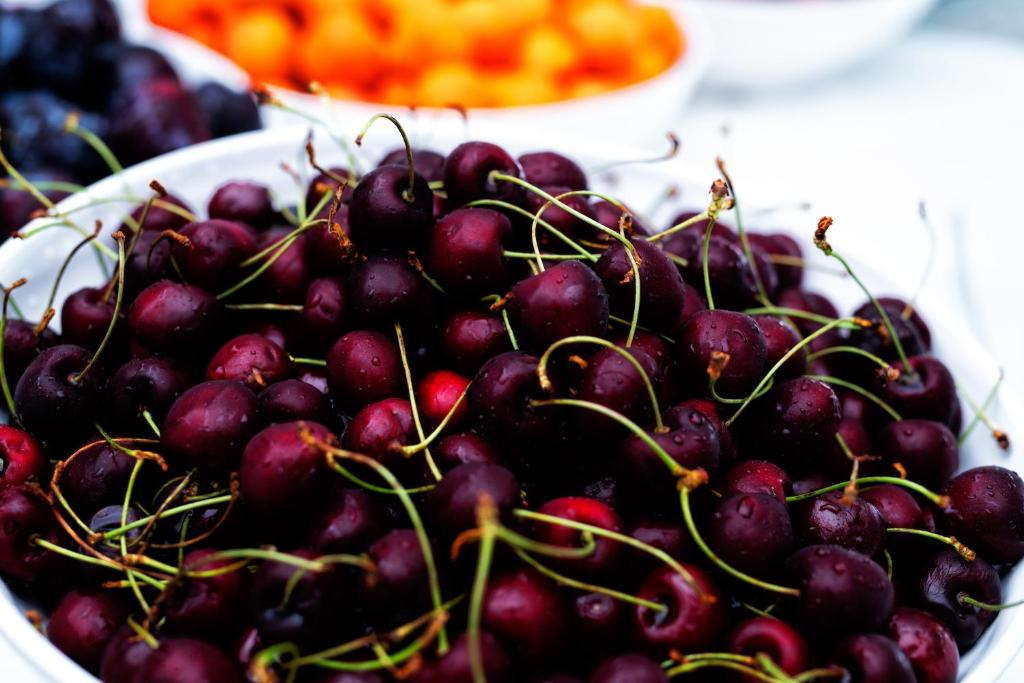 Melnās ķiršus kaudze ar spīdīgām miziņām un pilnām ogām, kas izkārtotas baltā traukā. Fons satur citus augļus, piešķirot dzīvīgu atmosfēru.