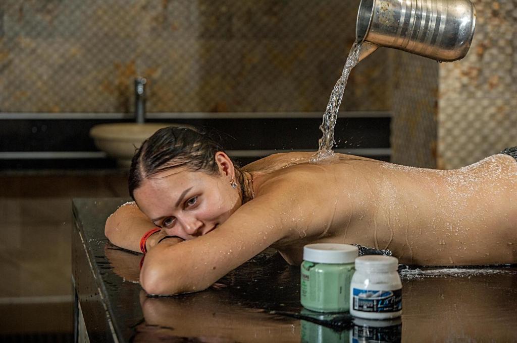 Sieviete guļ uz galda, kamēr ūdens plūst no metāla kannas uz viņas muguras. Blakus ir vairāki krēmi. Atmosfēra ir relaksējoša un spa stila.