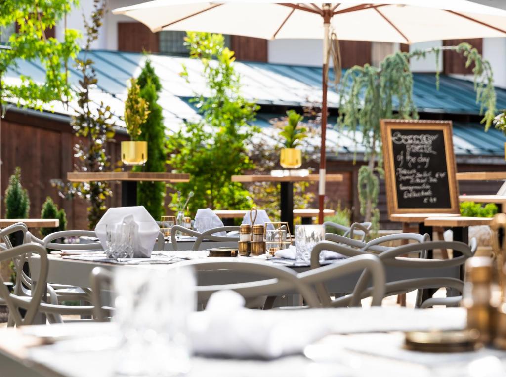 Āra restorāna terase ar galdiem, saulessargiem un zaļiem augiem, radot patīkamu un relaksējošu atmosfēru. Redzams melns tāfeles ēdienkarte ar piedāvājumiem.