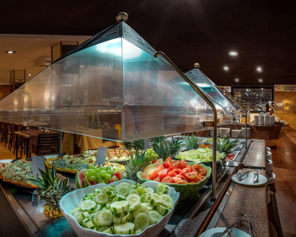Bufetes stends ar svaigiem dārzeņiem un augļiem, ieskaitot gurķus, tomātus un ananasus, apgaismots ar mūsdienīgu apgaismojumu. Fonā redzamas ēdināšanas vietas detaļas.