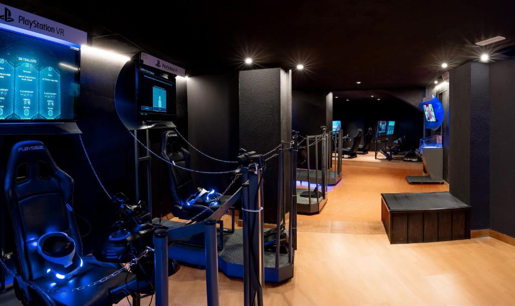 Virtuālās realitātes spēļu zona ar PlayStation VR, kurā redzamas ērtas spēļu sēdvietas un monitora ekrāni. Mūsdienīgs interjers ar zemu apgaismojumu un tehnoloģiskām ierīcēm.
