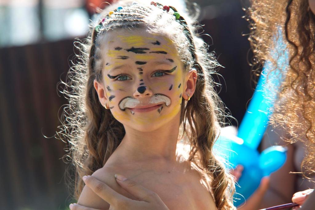 Bērns ar krāsainu sejas apgleznojumu, smaidošs saulainā dienā.