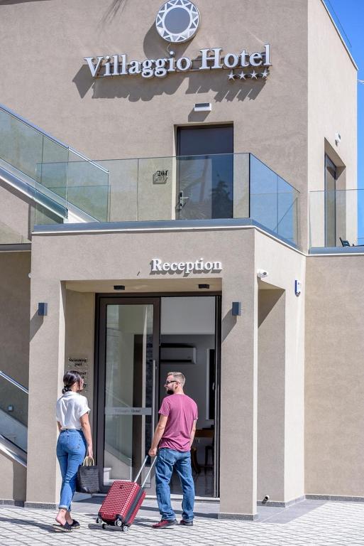 Villaggio Hotel ieeja ar modernu arhitektūru un skaidru "Reģistrācija" zīmi. Pāris ar bagāžu dodas uz viesnīcu, lai uzsāktu savu atpūtu.