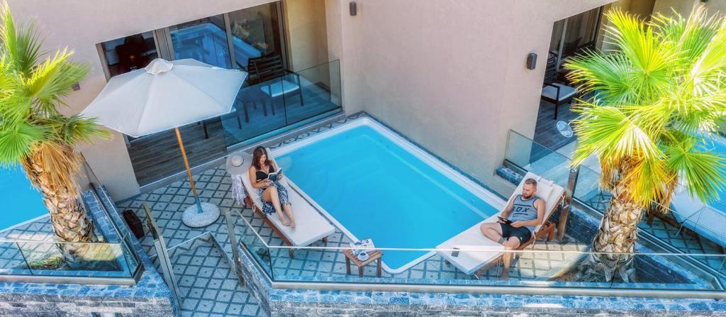 Atpūtas zona pie peldbaseina ar palmām un saulessargu, kur divi cilvēki relaksējas uz guļamkrēsliem. Ideāla vide, lai izbaudītu sauli un mieru.