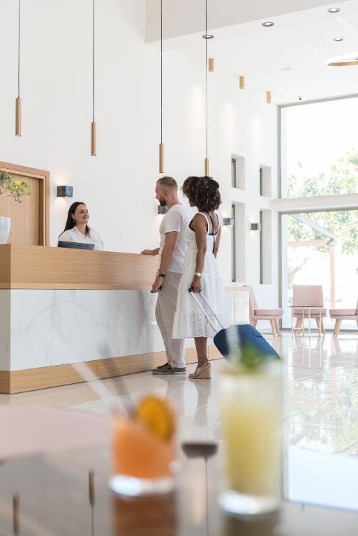 Recepcija modernā viesnīcā ar viesiem, kas reģistrējas, un laipnu darbinieku. Priekšplānā redzami kokteiļi.