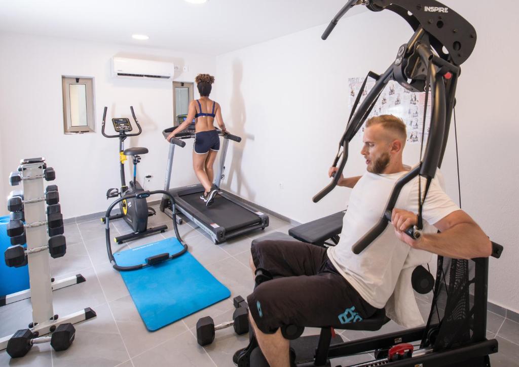 Trenažieru zāle ar diviem sportistiem, kas izmanto dažādas fitnesa iekārtas, piemēram, skrejceliņu un trenažieri. Iekārta ir moderna un telpa ir gaiša un plaša.