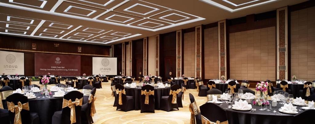 Eleganti iekārtota banketu zāle ar melniem galdiem, kas rotāti ar zelta lentēm. Zālē redzami ziedi un galda piederumi, radot svinīgu atmosfēru.