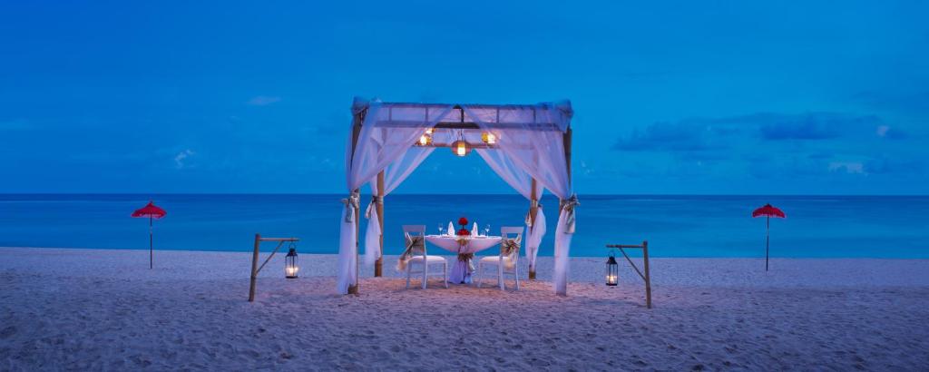 Romantiska vakariņas pludmalē zem balta nojumes ar siltu apgaismojumu un sarkaniem saulessargiem pie ūdens.