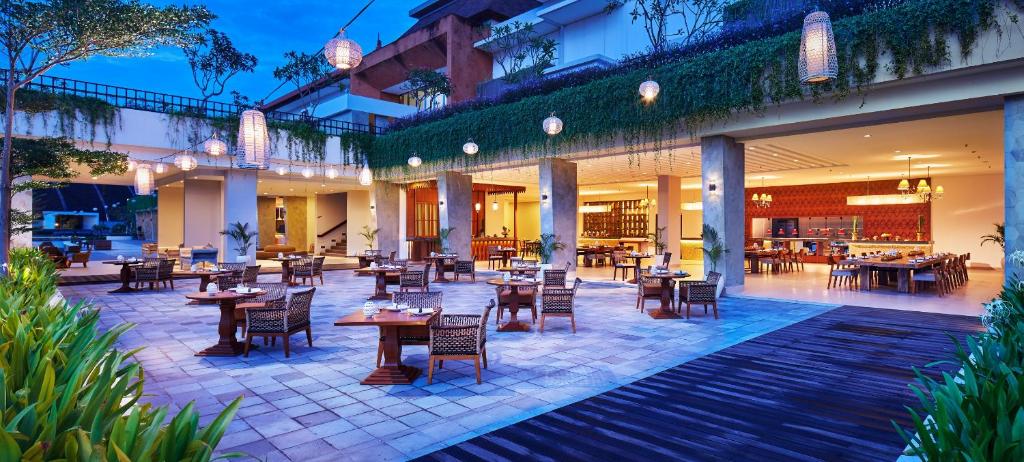 Eleganta āra restorāna zona ar sēdvietām, apgaismotām ar siltām lampām, kas radījušas patīkamu atmosfēru. Apkārtnē redzami zaļumi un mūsdienīgs arhitektūras dizains.