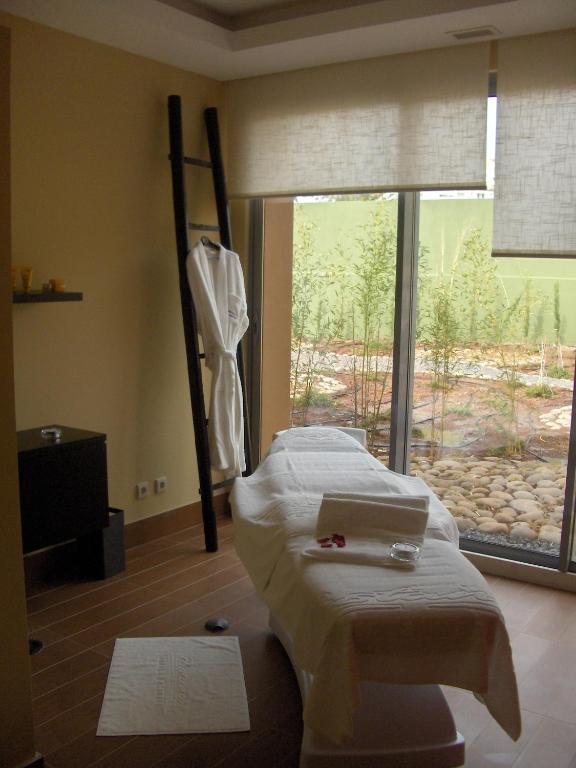 Relaksējoša spa telpa ar masāžas galdu, logiem, caur kuriem redzama dārza ainava, un baltiem halātiem. Ideāla vide atpūtai un labsajūtai.