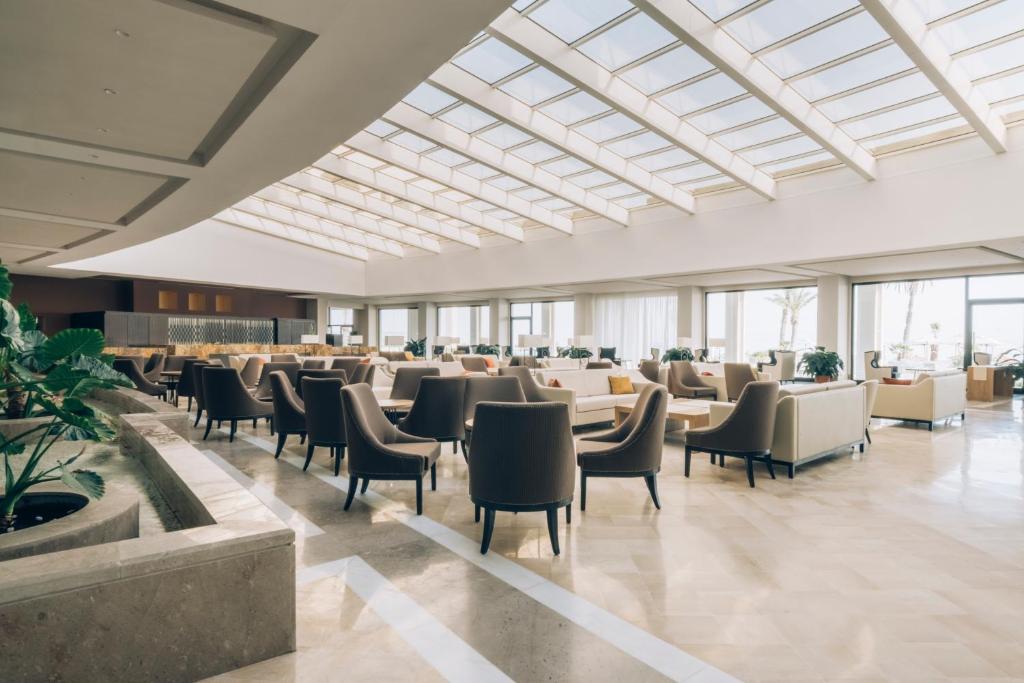 Moderns viesnīcas salons ar plašām telpām, lieliem logiem un elegantiem sēdekļiem, kas nodrošina komfortablu atmosfēru. Gaismas plūsma caur stikla jumtu rada patīkamu un atvērtu vidi.