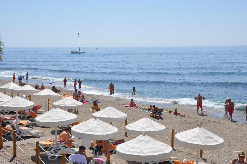 Pludmales ainava ar saulainām dienām, kur cilvēki atpūšas zem saulessargiem, peldas jūrā un baudā saulainu laiku. Fons ar jachtu un mierīgu ūdeni papildina pludmales atmosfēru.