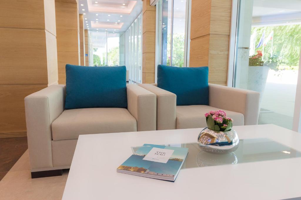 Mūsdienīgs atpūtas stūris ar divām gaišām krēslām un ziliem spilveniem, baltu galdu un dekoratīvu podiņu ar ziediem. Interjers ir gaišs un elegants, radot patīkamu atmosfēru.