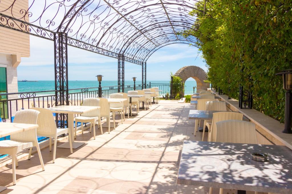 Skaists restorāna terases skats ar baltiem galdiem un krēsliem, kas vērsti uz jūru, zem izsmalcināta metāla nojumes. Lieliska vieta relaksācijai ar dabu un ūdeni fonā.