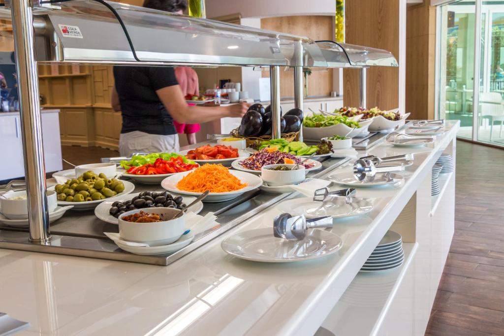 Bagātīgs salātu bufete ar dažādiem dārzeņiem un piedevām, ieskaitot olīvas, tomātus un rīvētas burkānus, viesnīcā. Dabiski apgaismotā telpā redzami arī trauki un galda piederumi.