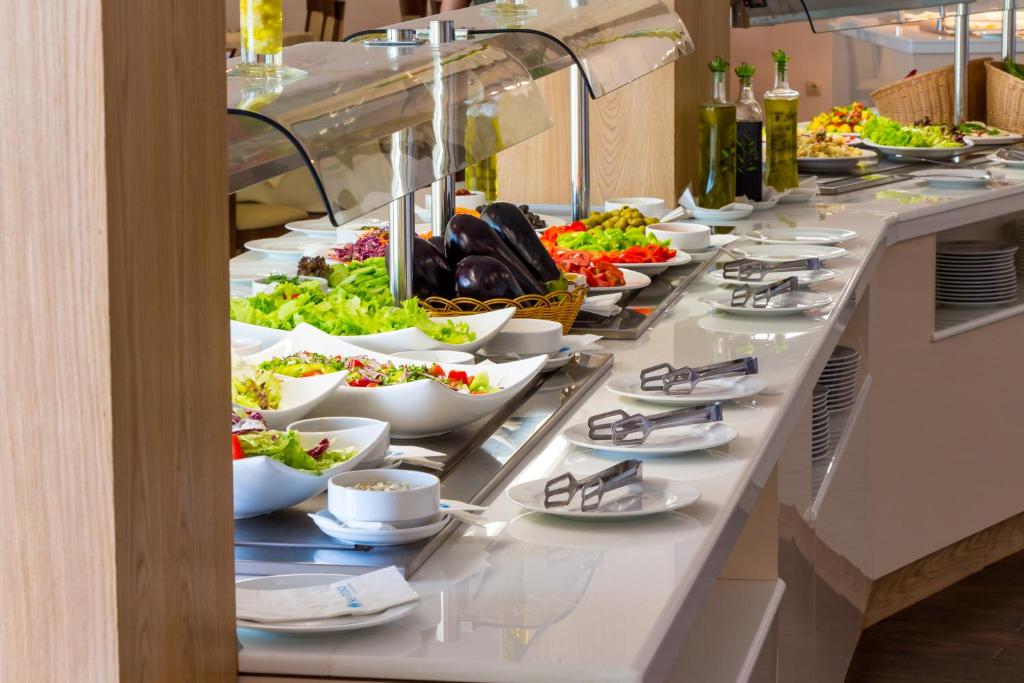 Bagātīgs salātu bufete ar dažādām dārzeņu un zaļumu izvēlēm, kas pasniegta baltos traukos. Galda pārklājums ar ēdiena pasniegšanai un dekoratīviem elementiem.