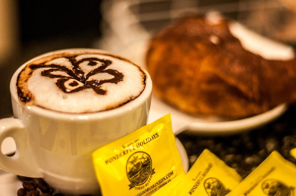 Kafijas krūze ar mākslinieciski veidotu putu zīmējumu, blakus ir saldā maize un cukura paciņas.