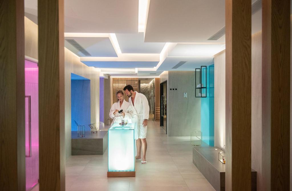 Mūsdienīga spa telpa ar apgaismojumu, kurā divi cilvēki baltos halātos apspriež procedūras. Svaiguma un relaksācijas atmosfēra.