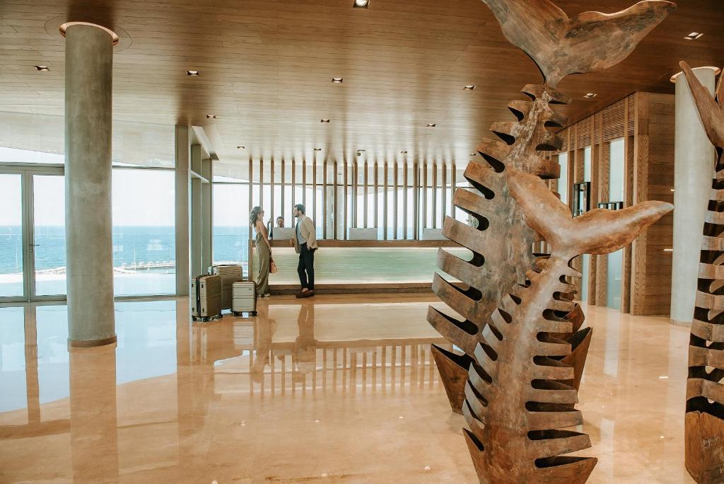 Mūsdienīgs viesnīcas interjers ar jūras skatu, kurā redzami divi ceļotāji pie reģistratūras un unikālas zivs formas skulptūras.