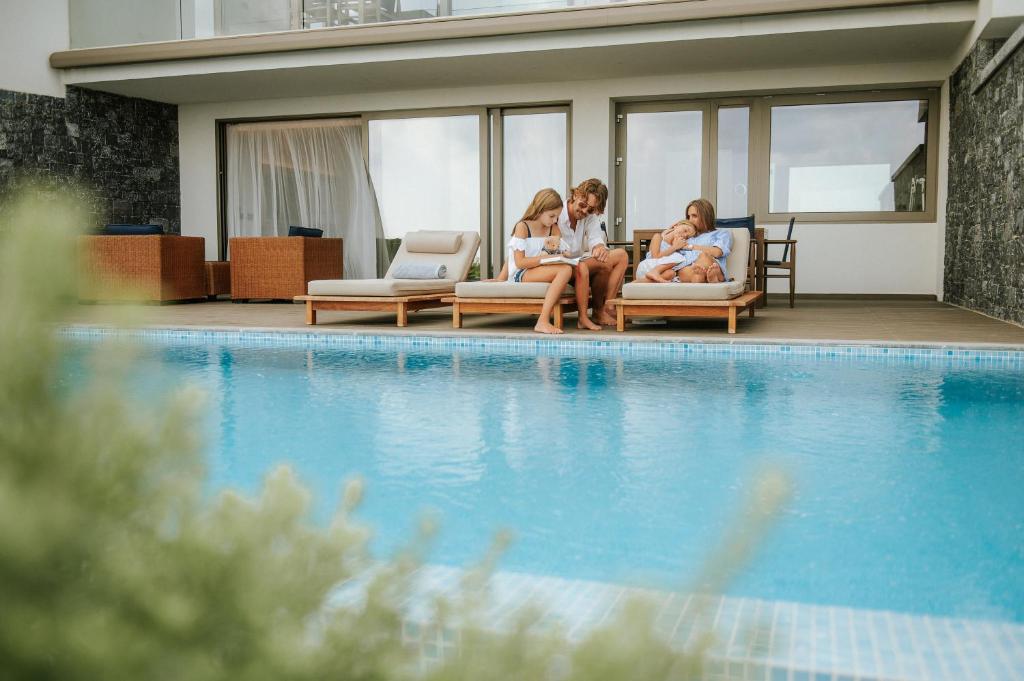 Ģimene atpūšas blakus baseinam, izbaudot laiku kopā, lasot un relaksējoties. Mūsdienīgs interjers un skaidrs laiks veido patīkamu atmosfēru.