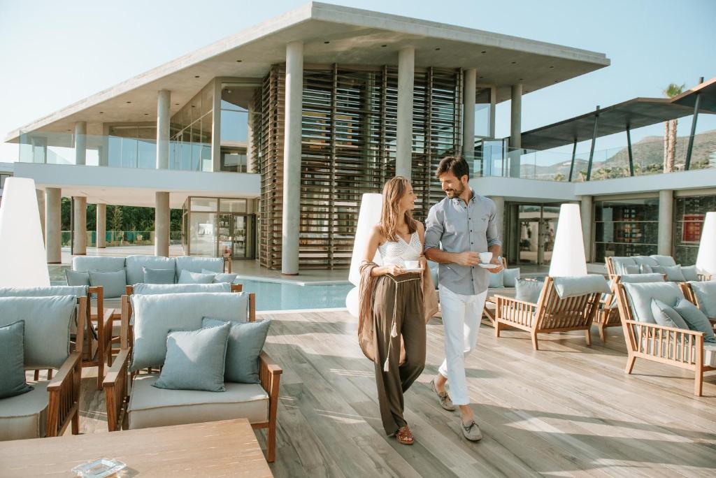Pāris pastaigājas pa modernas viesnīcas terasi, baudot kafiju un saulainu laiku. Apkārt ir eleganti mēbelēti sēdvietas un baseins, kas veido relaksējošu atmosfēru.