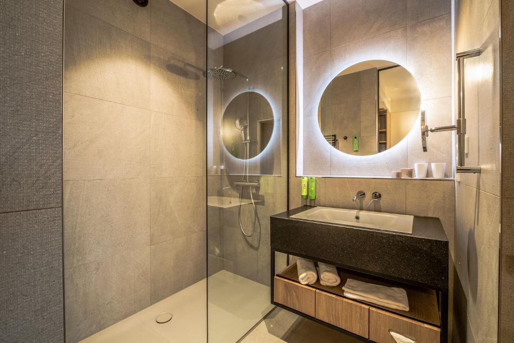 Mūsdienīgs vannas istabas dizains ar stikla dušu un melnu izlietni, kas papildināta ar apgaismojumu. Interjers ir veidots ar pelēku flīžu un koka elementu kombināciju.