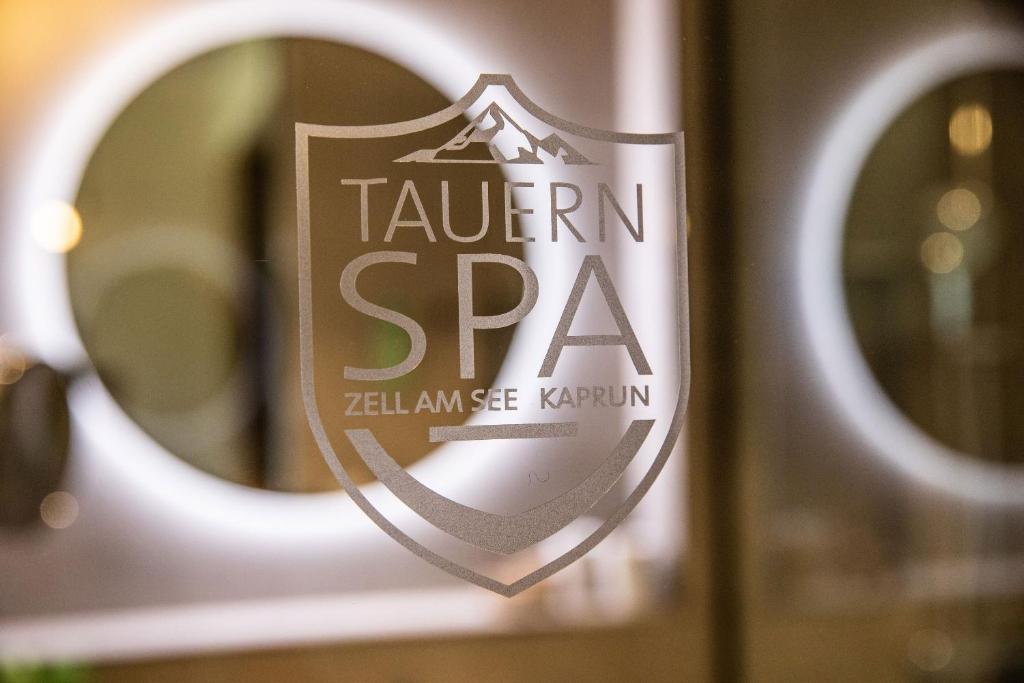 Tauern SPA logo uz stikla virsmas, ar apakšā norādītu "Zell am See Kaprun". Elegants un moderns dizains, kas atspoguļo relaksācijas atmosfēru.