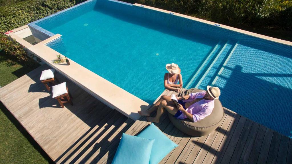 Divi cilvēki atpūšas pie baseina, baudot sauli un relaksāciju. Apkārt ir koka terase un zaļumi, radot mierīgu atmosfēru.