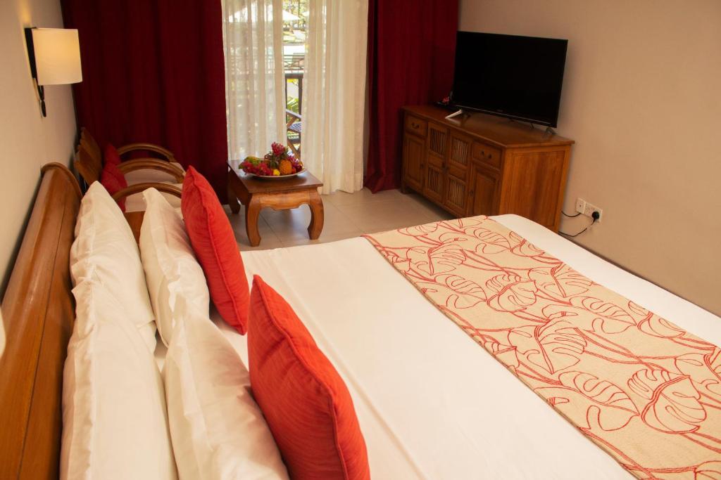 Mūsdienīga viesnīcas istaba ar plašu gultu, spilveniem un sarkaniem akcentiem. Istabā ir koka mēbeles, televizors un dabiska apgaismojuma no loga.