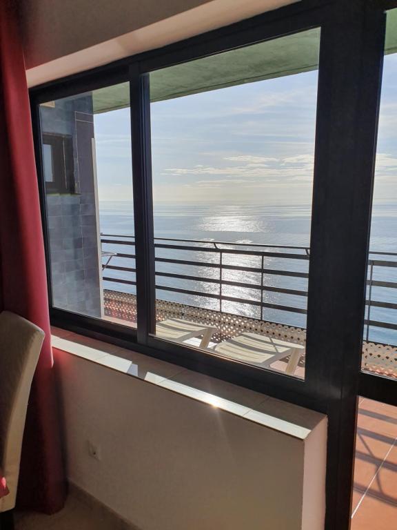 Skats no viesnīcas numura uz jūru caur lielu logu, ar terasi un saule, kas atspīd uz ūdens.