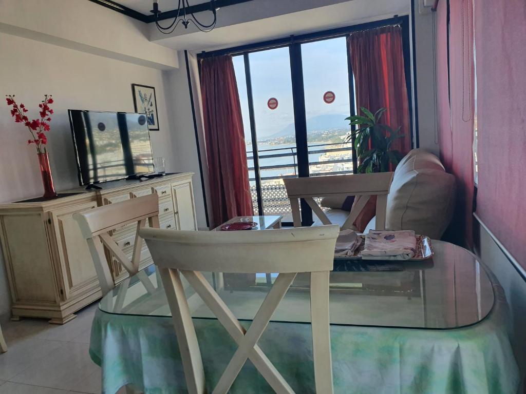 Mūsdienīga dzīvojamā istaba ar panorāmas logiem, televizoru un ēdamgaldu. Interjerā ir sarkanas aizkari un augi, radot mājīgu atmosfēru.