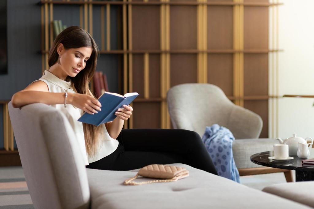 Sieviete, kas lasa grāmatu komfortablā interjerā, ar tēju un dekoriem blakus. Rāmā atmosfērā izceļas viņas koncentrācija un relaksācija.