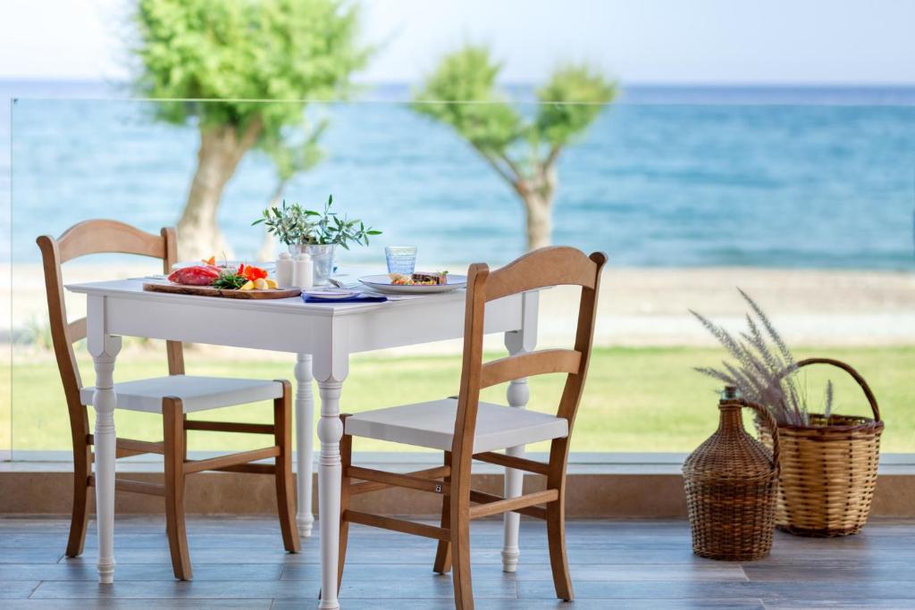 Skaista ēdamistaba ar baltu galdu un koka krēsliem, kurā redzama garša pārtika, ar skatu uz jūru un zaļām koku lapām. Ideāla vieta relaksējošām brokastīm pie ūdens.