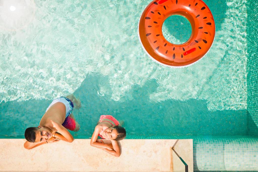 Divi bērni peld baseinā, priecājoties un atpūšoties, ar oranžu ūdens gredzenu virsū. Ūdens ir dzidrs un spožs, radot vasarīgu atmosfēru.