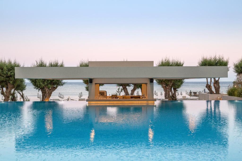 Mūsdienīgs ūdens baseins ar elegantu atpūtas zonu, kas atrodas pie jūras ar zilu ūdeni un zaļiem kokiem fonā. Ideāla vieta relaksācijai un mierīgām brīvdienām.