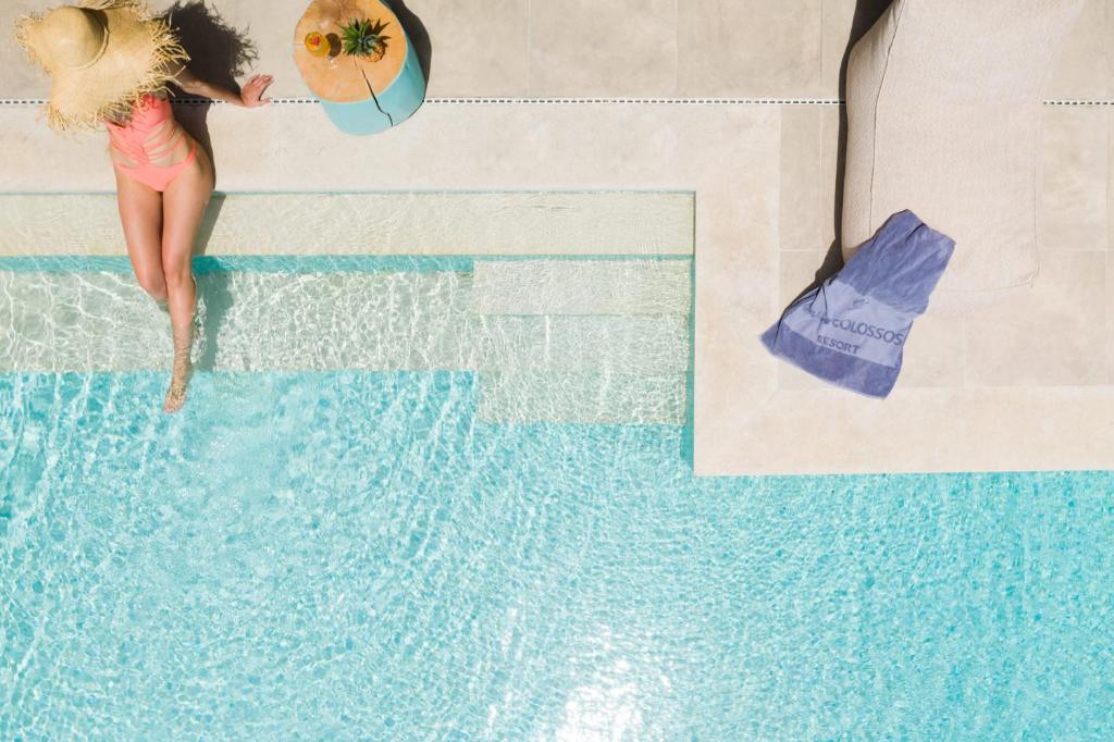 Sieviete rozā peldkostīmā atpūšas pie baseina, valkājot lielu saules cepuri. Blakus ir krēsls un dvielis, uz kura redzama "Golosso Resort" logo.