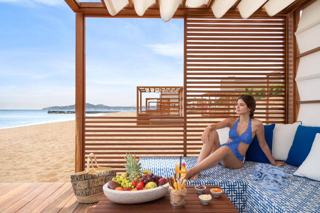 Pludmales atpūtas zona ar modernu koka struktūru, kurā ir sēžamvieta un augļu grozs. Viegla, saulaina atmosfēra un mierīga jūra fonā.