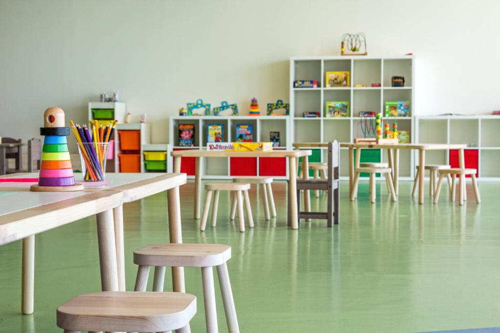 Krāsains bērnudārza interjers ar koka galdiem un krēsliem, rotaļlietām un krāsu zīmuļiem. Telpai ir gaiša un draudzīga atmosfēra, kas veicina radošumu un mācīšanos.
