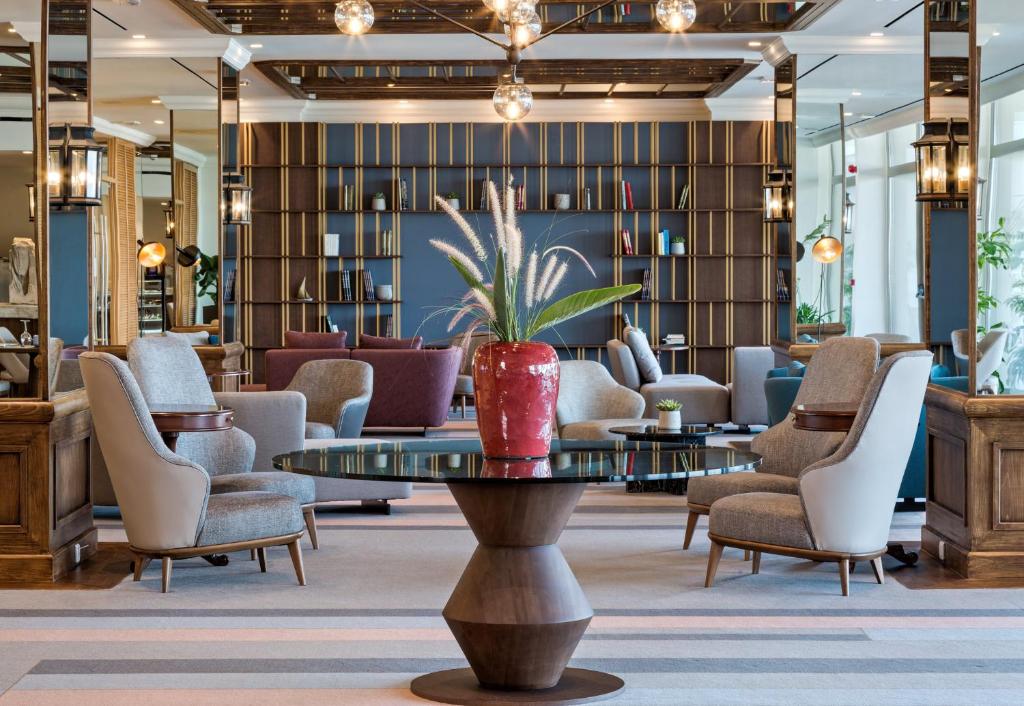 Mūsdienīgs viesnīcas interjers ar elegantiem krēsliem, stikla galdu un dekoratīvām augu kompozīcijām. Siltas krāsas un apgaismojums rada patīkamu atmosfēru.