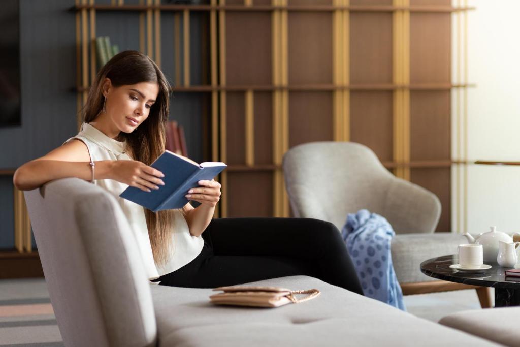Sieviete, kas lasīja grāmatu komfortablā interjerā, sēžot uz dīvāna ar tējas krūzīti blakus. Rāmā atmosfērā redzamas modernas mēbeles un grāmatu plaukti.