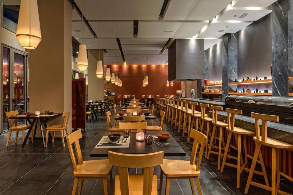 Mūsdienīgs restorāna interjers ar koka mēbelēm un maigu apgaismojumu, kas rada siltu atmosfēru. Galdi ir sakārtoti, gatavi viesiem, un redzama sushi bārs.