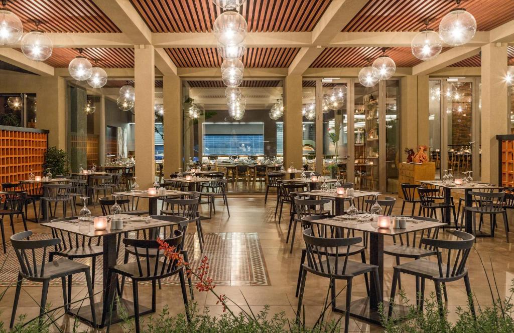 Mūsdienīgs restorāns ar elegantiem galdiem un krēsliem, ko apgaismo stilīgas gaismas. Pateicoties atvērtajiem logiem un zaļajiem augiem, telpa izskatās gaiša un aicinoša.