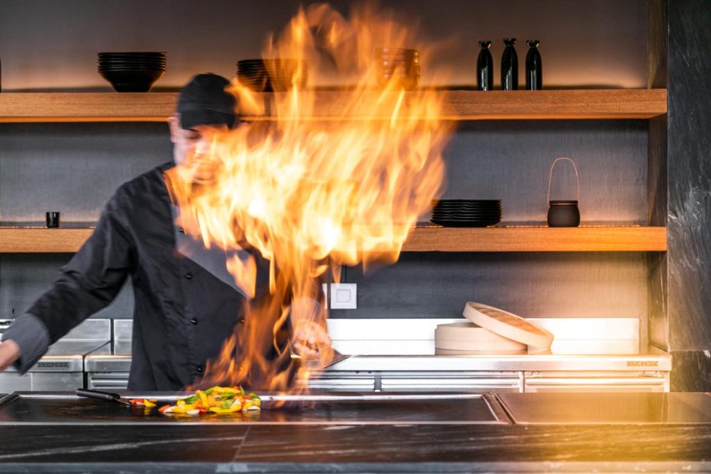 Kāds šefpavārs gatavo ēdienu, radot aizraujošu liesmu, demonstrējot kulinārijas prasmes. Aiz fona redzama moderna virtuve ar koka plauktiem un traukiem.