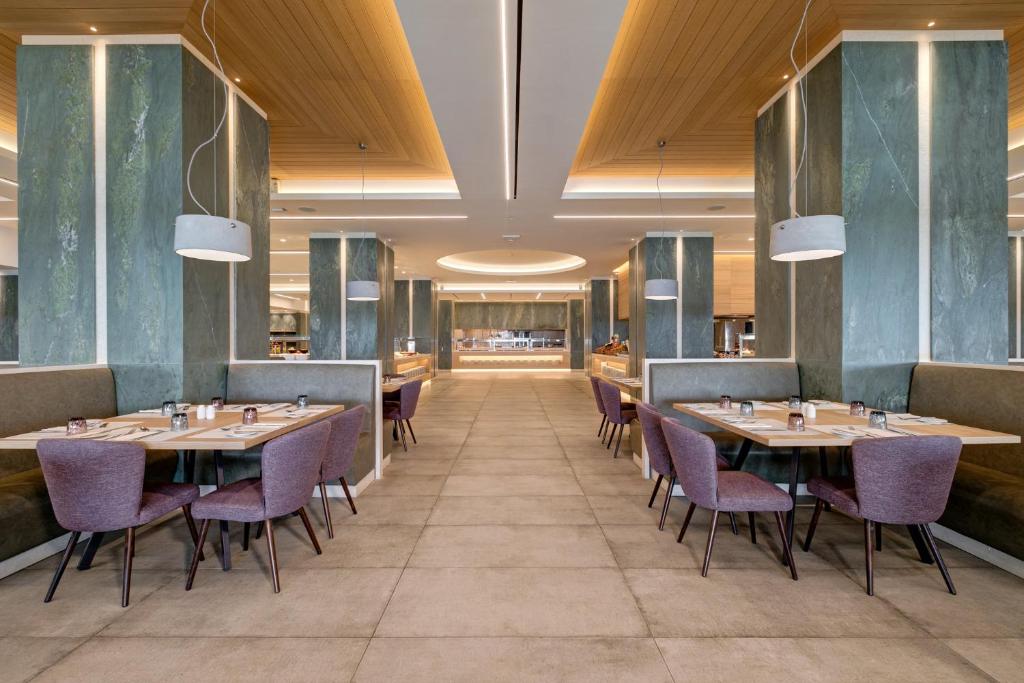 Mūsdienīgs restorāns ar elegantu interjeru, kurā redzami zaļi akmens stabi un stilīgas purpura krāsas krēsli pie koka galdiem. Apgaismojums rada siltu atmosfēru, izceļot telpas plašumu un komfortu.